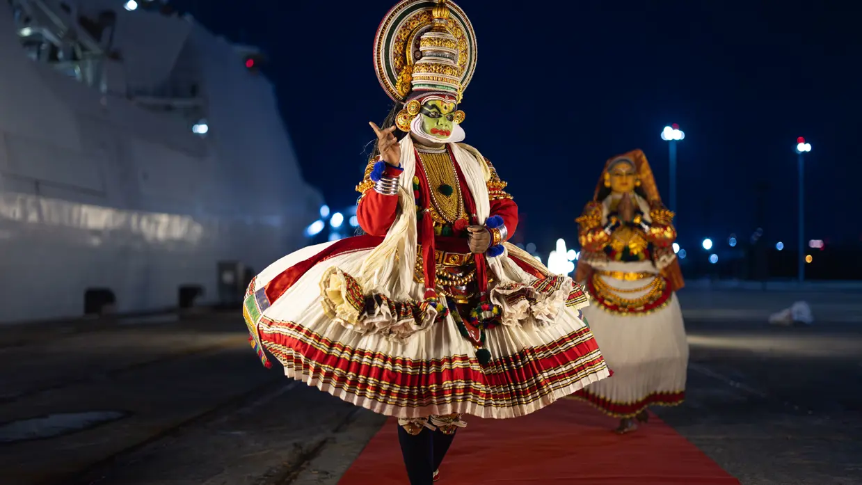 Een traditioneel geklede en gemaskerde Indiër op een rode loper naast de De Ruyter.  &nbsp;