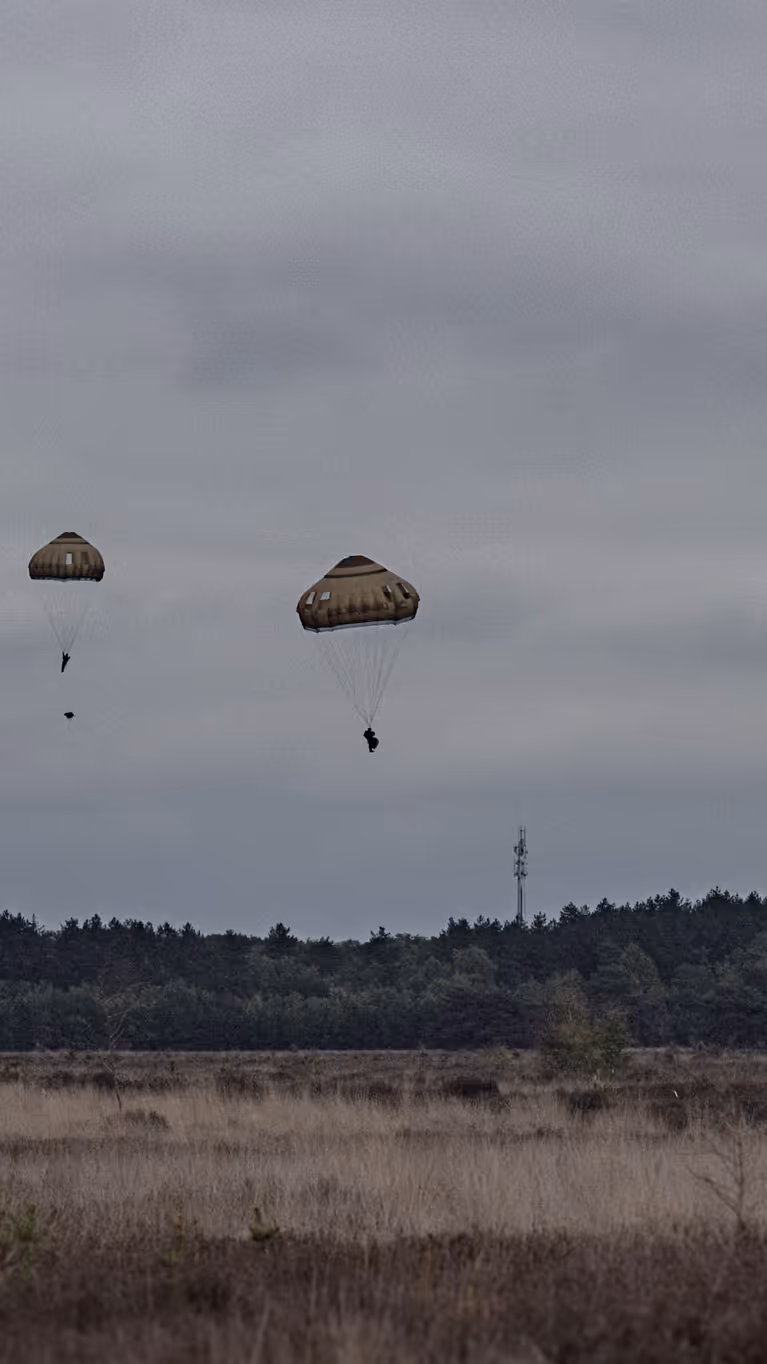 Meerdere parachutisten dalen neer boven de Belgische heide terwijl een transportvliegtuig op de achtergrond wegvliegt tijdens oefening Versatile Guard.