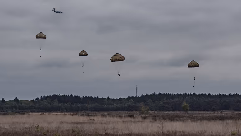 Meerdere parachutisten dalen neer boven de Belgische heide terwijl een transportvliegtuig op de achtergrond wegvliegt tijdens oefening Versatile Guard.