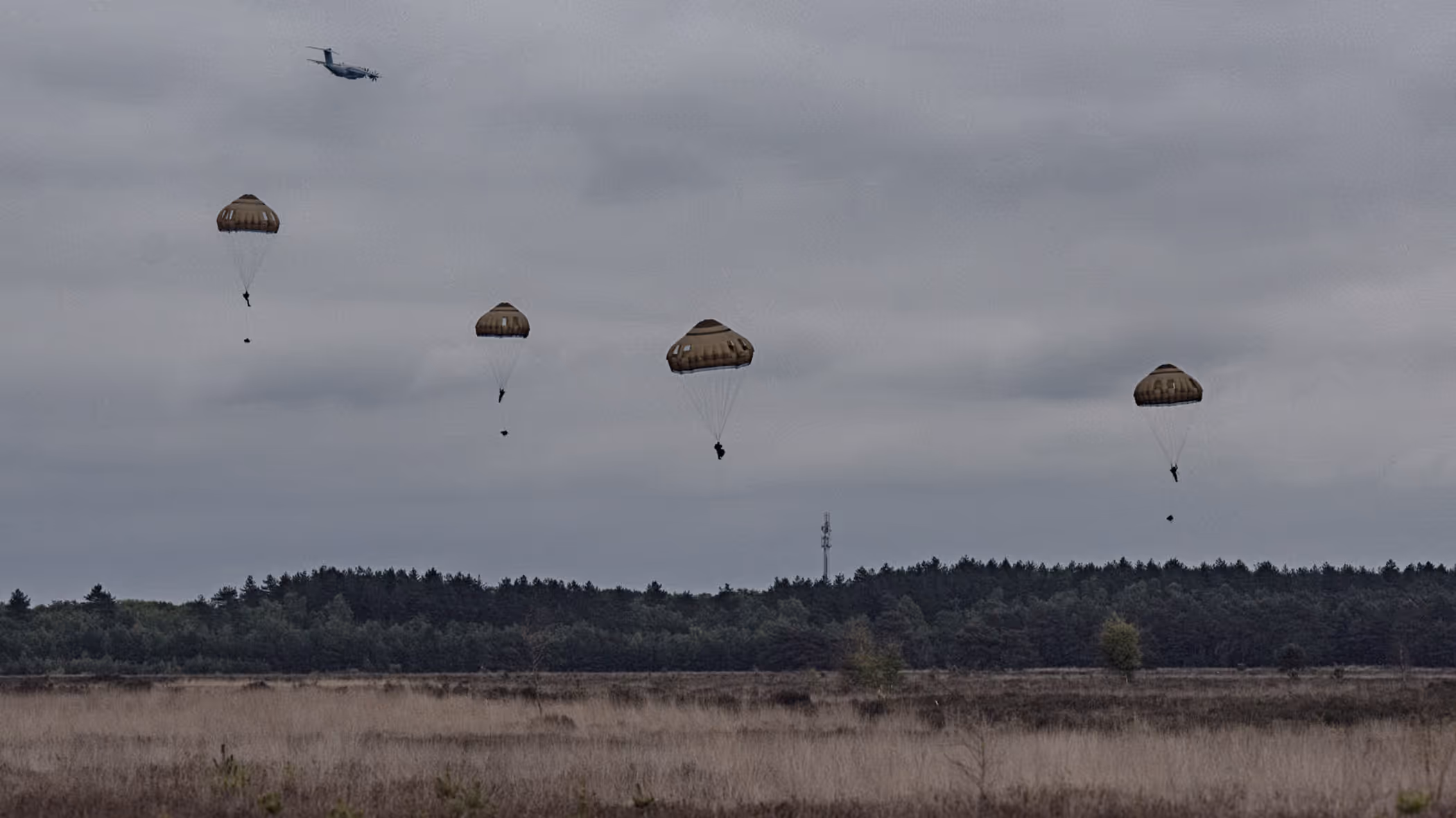 Meerdere parachutisten dalen neer boven de Belgische heide terwijl een transportvliegtuig op de achtergrond wegvliegt tijdens oefening Versatile Guard.