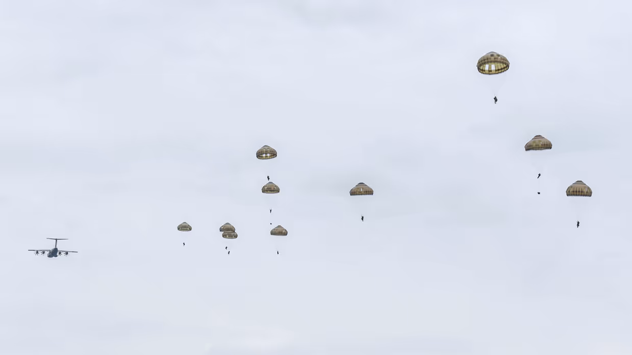 Tien parachutisten in de lucht met op de achtergrond een wegvliegend transportvliegtuig.