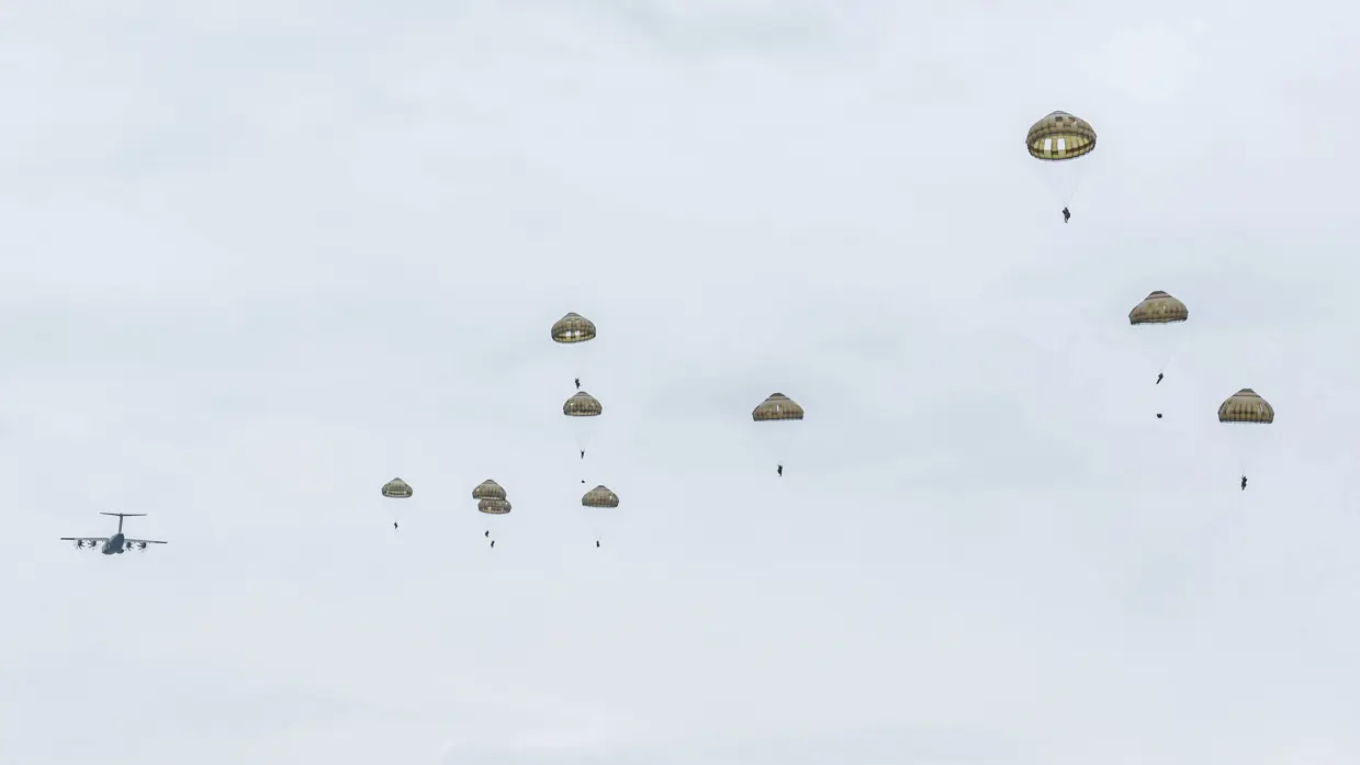 Tien parachutisten in de lucht met op de achtergrond een wegvliegend transportvliegtuig.