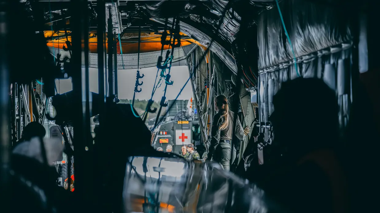 Vanuit de romp van de helikopter kijk je over materieel en een militair naar buiten door de openstaande laadklep. Daar staan militairen en een groen voertuig met daarop een rood kruis.