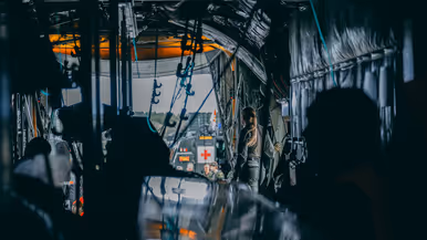 Vanuit de romp van de helikopter kijk je over materieel en een militair naar buiten door de openstaande laadklep. Daar staan militairen en een groen voertuig met daarop een rood kruis.