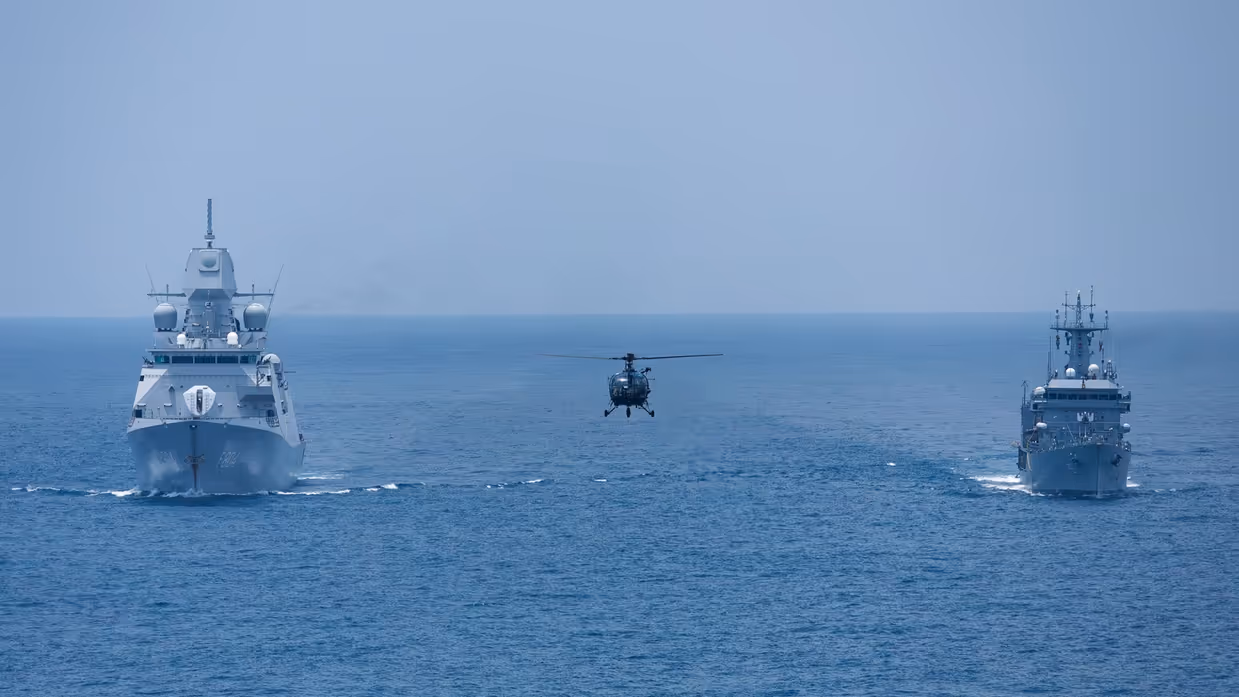 Tussen 2 marineschepen op zee vliegt een helikopter laag boven het water.