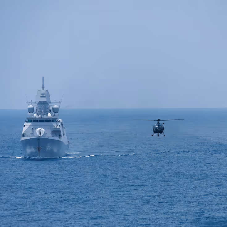 Tussen 2 marineschepen op zee vliegt een helikopter laag boven het water.