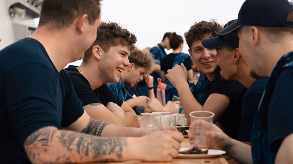 Lachende, jonge bemanningsleden zitten met een glaasje prik aan een campingtafel op het helidek.