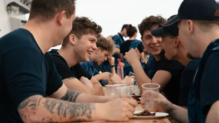 Lachende, jonge bemanningsleden zitten met een glaasje prik aan een campingtafel op het helidek.
