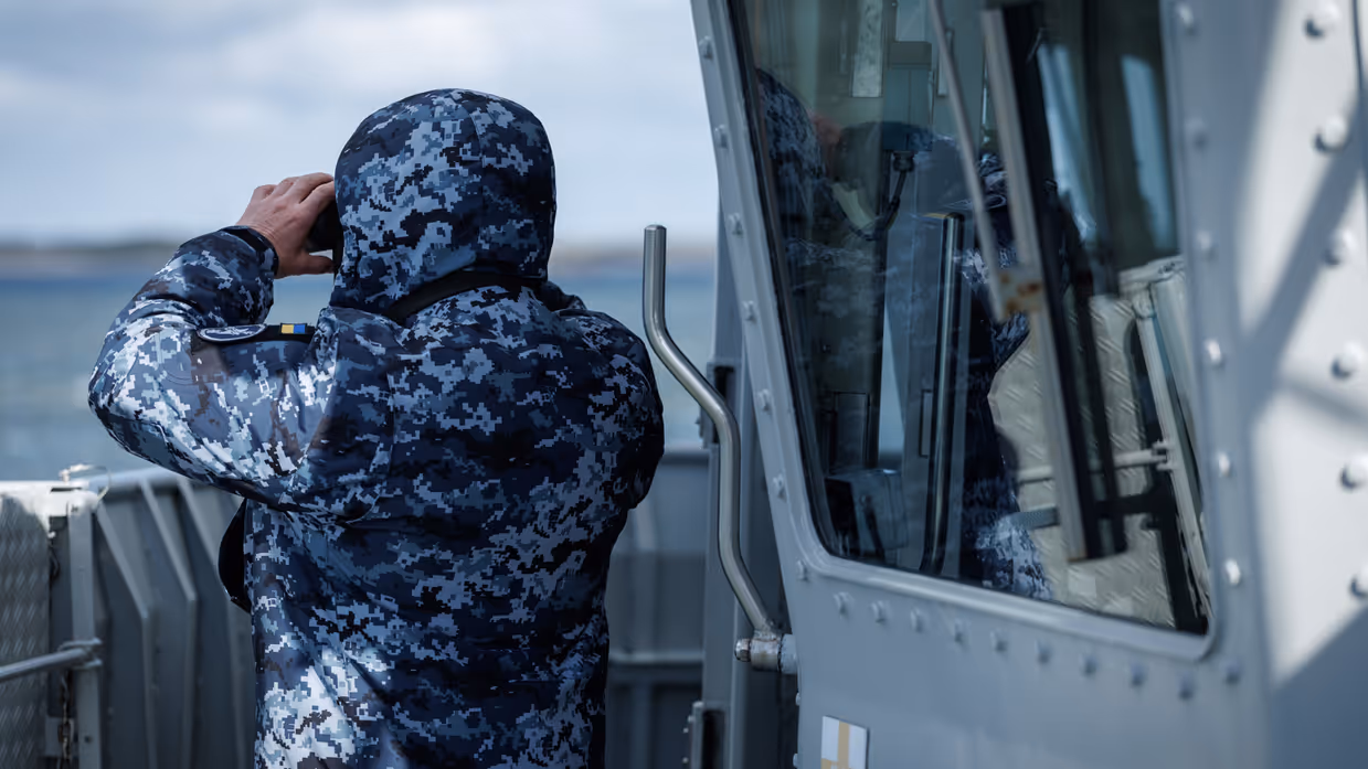 Een militair op de brug, een militair kijkt aan dek door een verrekijker en een militair bedient apparatuur op de brug.