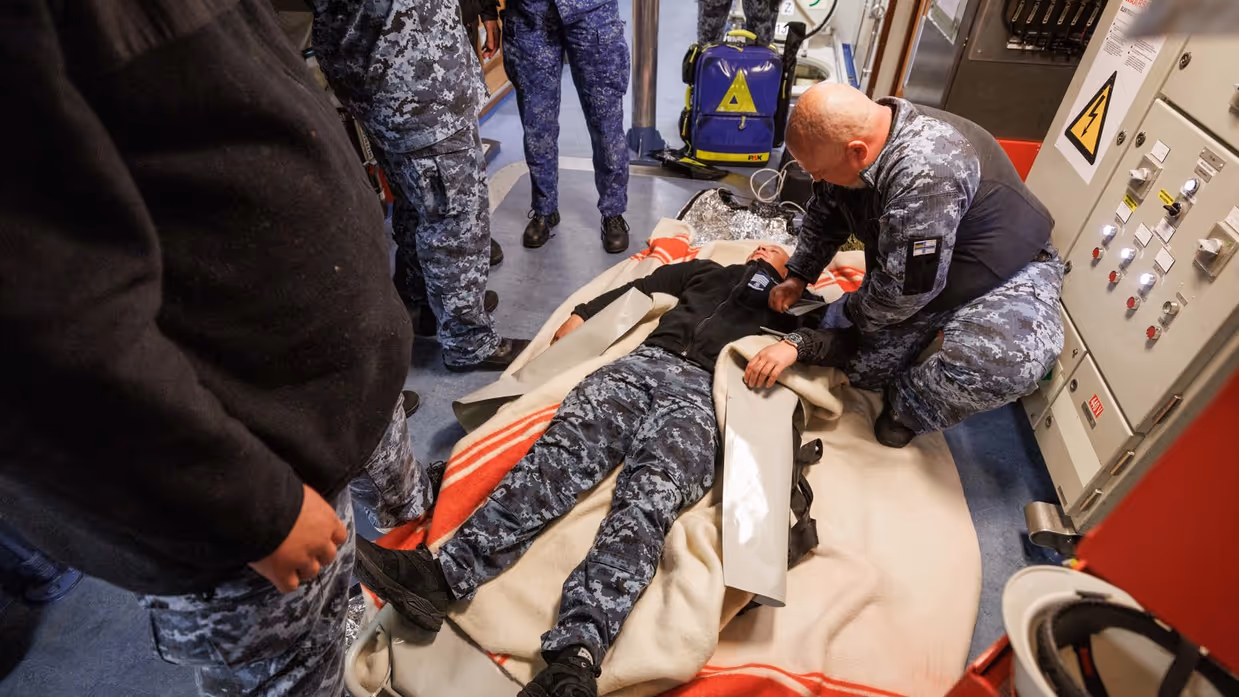 Militairen pakken tijdens de oefening de collega in die drenkeling speelt en warm moet worden gehouden en militairen behandelen deze oefengewonde in het schip.