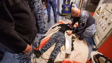 Militairen pakken tijdens de oefening de collega in die drenkeling speelt en warm moet worden gehouden en militairen behandelen deze oefengewonde in het schip.