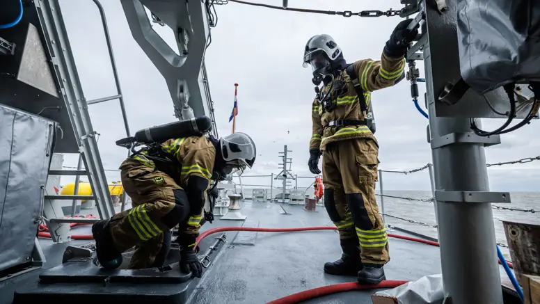 2 brandweermannen in geel pak werken op een marineschip, omringd door metalen constructies en een grijze zee.