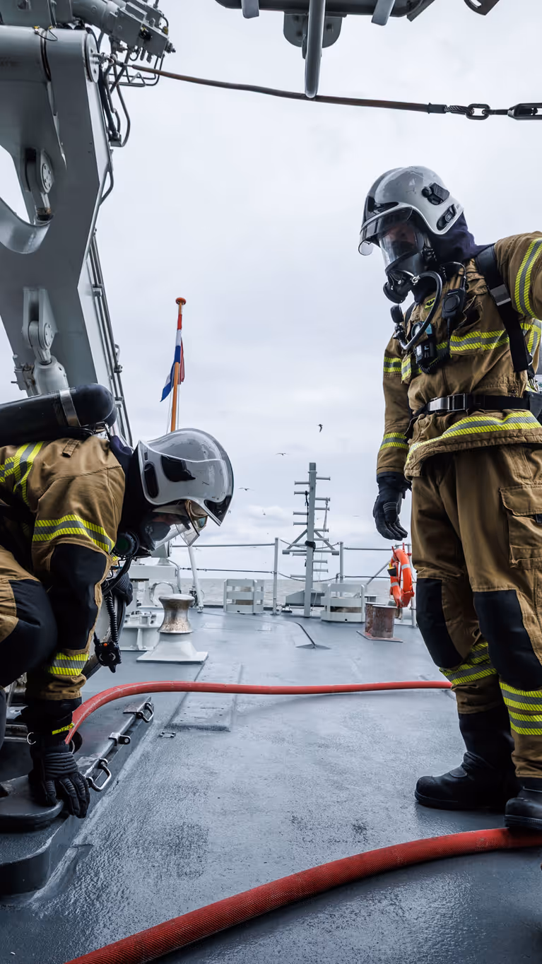 2 brandweermannen in geel pak werken op een marineschip, omringd door metalen constructies en een grijze zee.
