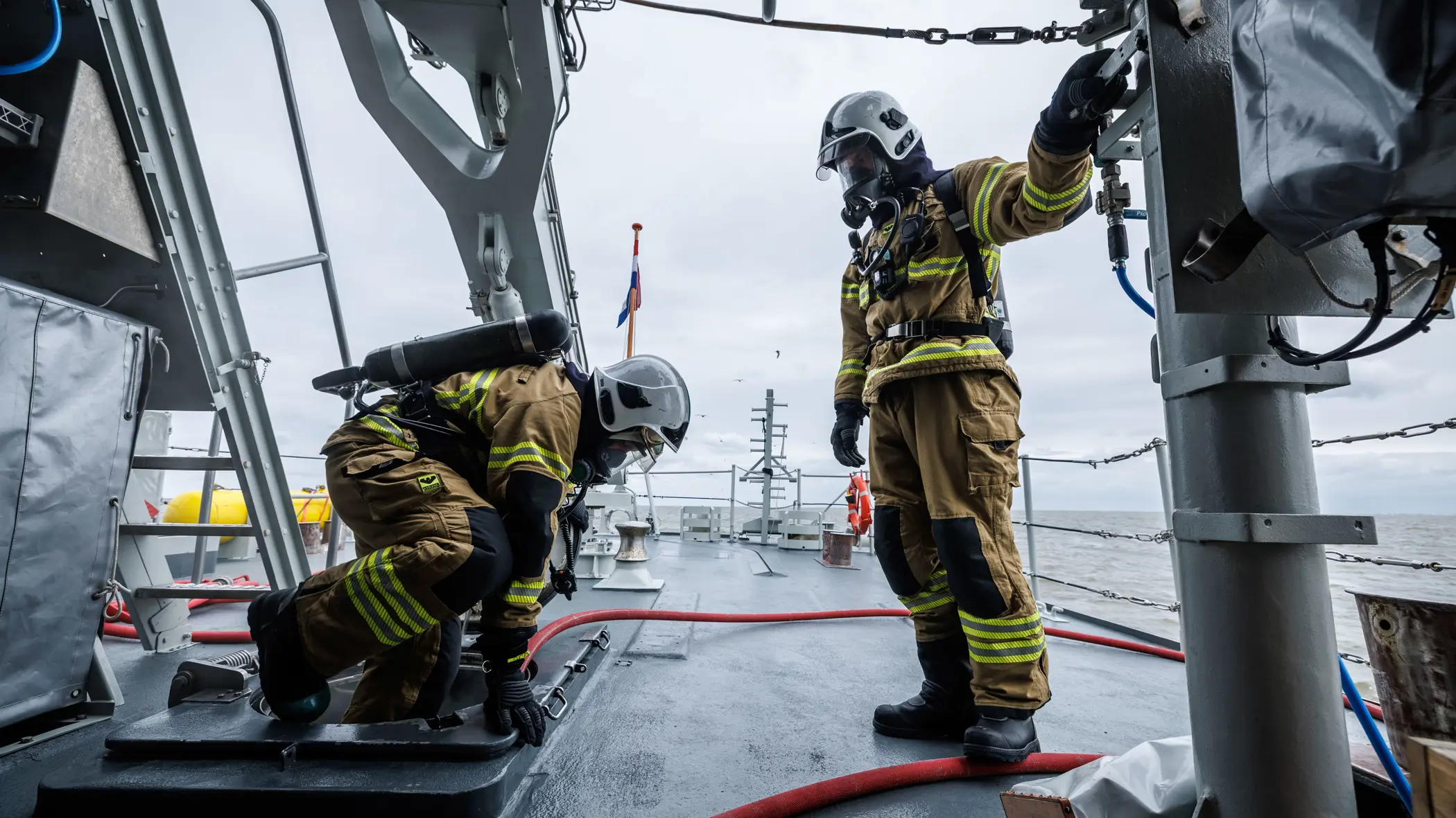 2 brandweermannen in geel pak werken op een marineschip, omringd door metalen constructies en een grijze zee.
