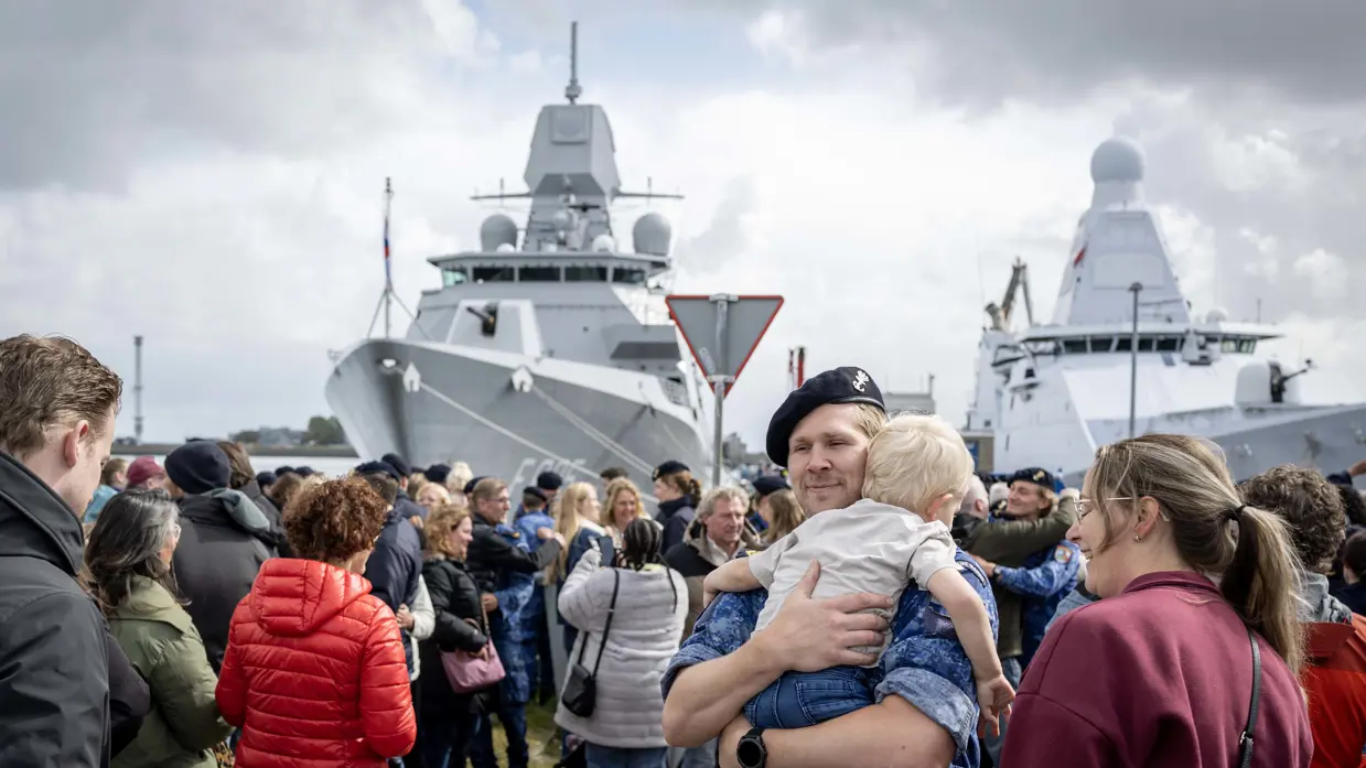 Een bemanningslid draagt een klein kind op zijn arm, omringd door anderen, met twee schepen op de achtergrond.