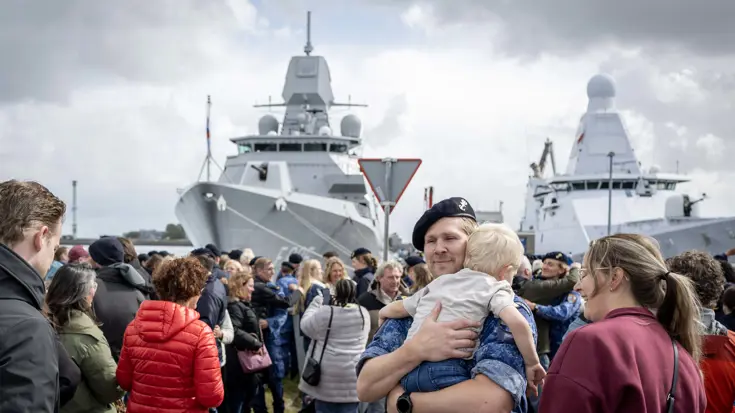 Een bemanningslid draagt een klein kind op zijn arm, omringd door anderen, met twee schepen op de achtergrond.