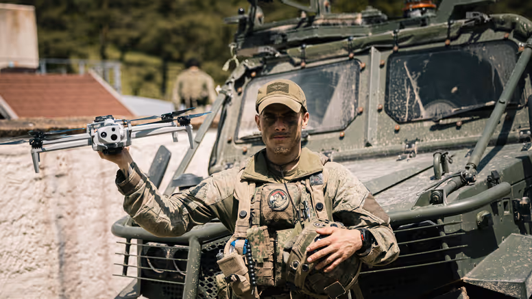 Een militair van de bevoorradingscompagnie van de Luchtmobiele Brigade voor een voertuig met een drone in zijn hand.
