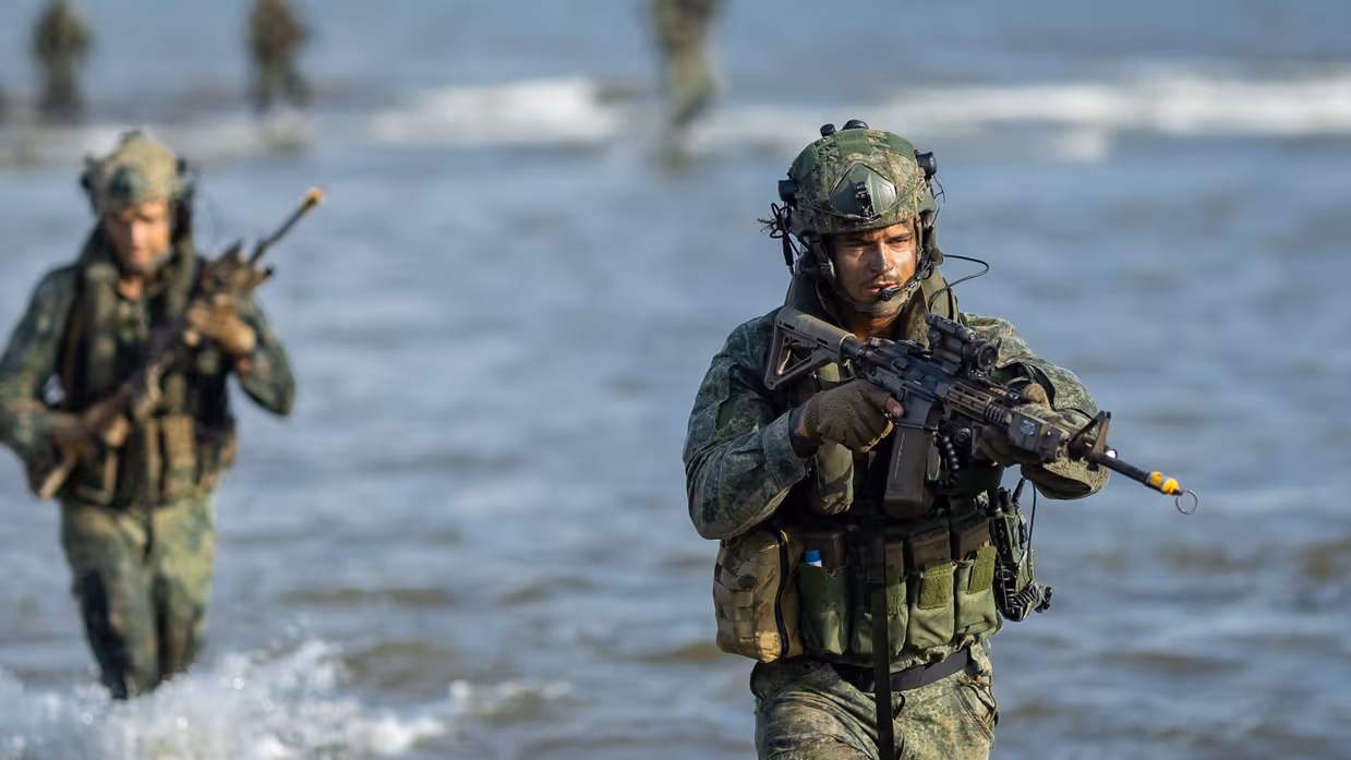 Een marinier loopt door het water, mariniers op het strand, mariniers verdekt opgesteld en een marinier roept bevelen.
