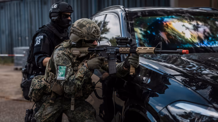 Militair richt zijn wapen op een auto. Daarachter staat iemand van de BE.