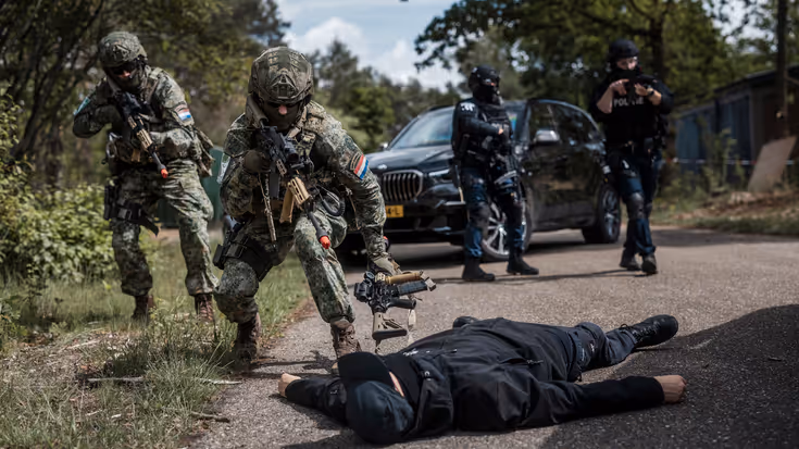 Op de voorgrond een acteur-terrorist die overmeesterd is met 2 militairen met wapen in de aanslag.