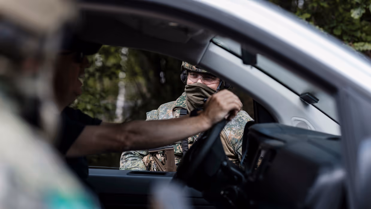 Een man in een voertuig, waar een militair een gesprek mee voert door het voorraam van dit voertuig.
