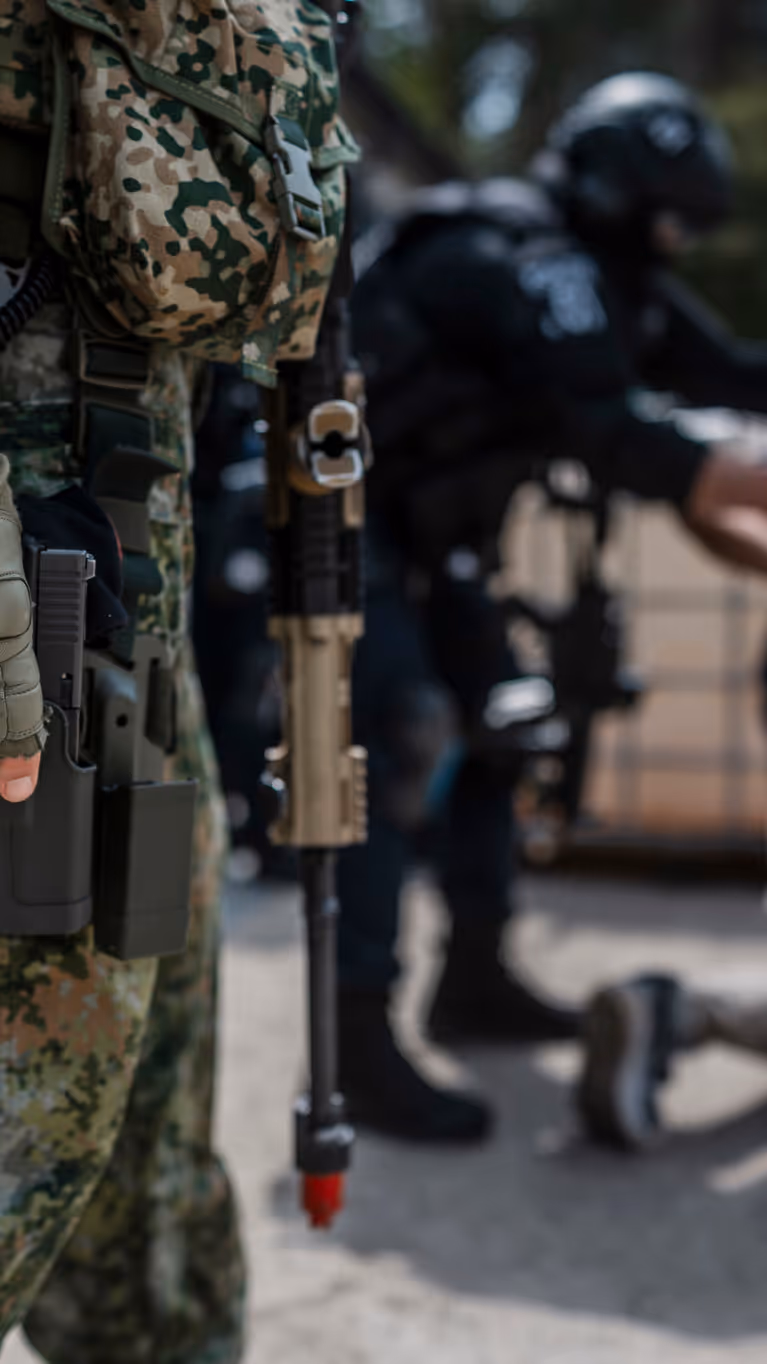 Militair staat op de voorgrond paraat met wapen en op de achtergrond wordt iemand aangehouden door de Politie.