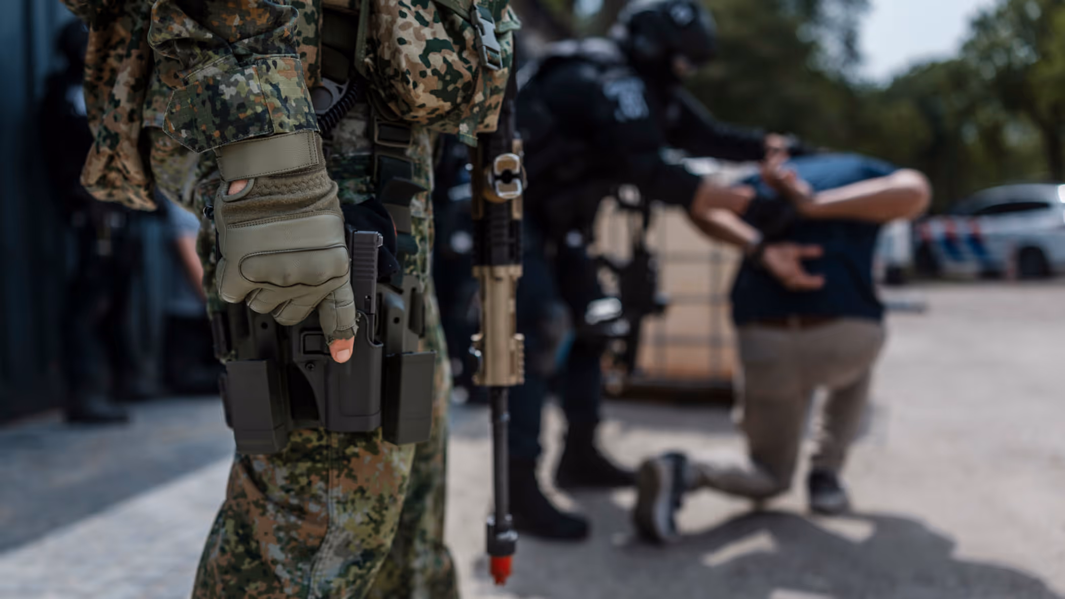 Militair staat op de voorgrond paraat met wapen en op de achtergrond wordt iemand aangehouden door de Politie.