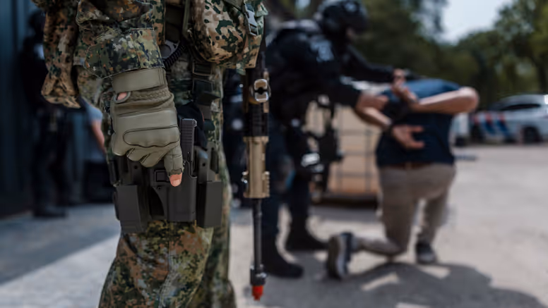 Militair staat op de voorgrond paraat met wapen en op de achtergrond wordt iemand aangehouden door de Politie.