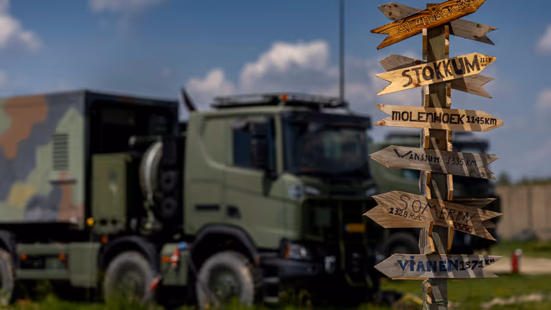 Een paal met daarop allemaal wegwijzers naar plaatsen in Nederland inclusief de afstand. Op de achtergrond staat een vrachtwagen.