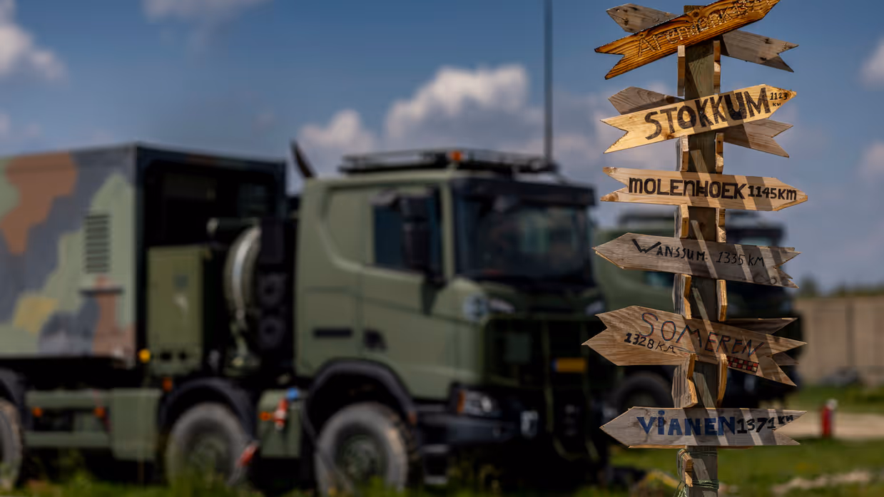 Een paal met daarop allemaal wegwijzers naar plaatsen in Nederland inclusief de afstand. Op de achtergrond staat een vrachtwagen.
