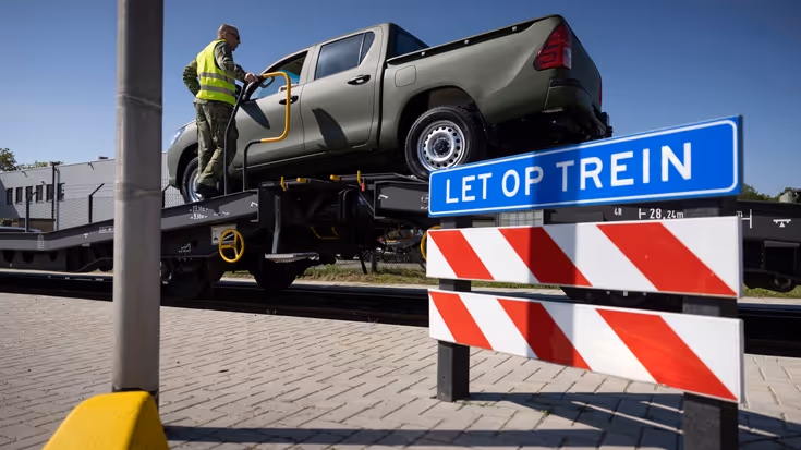 Een pick-up truck staat op een goederenwagon, ernaast staat een militair in geel hesje. Op de voorgrond staat een rood-wit bord met erboven een blauw bord met de tekst ‘let op trein’. 