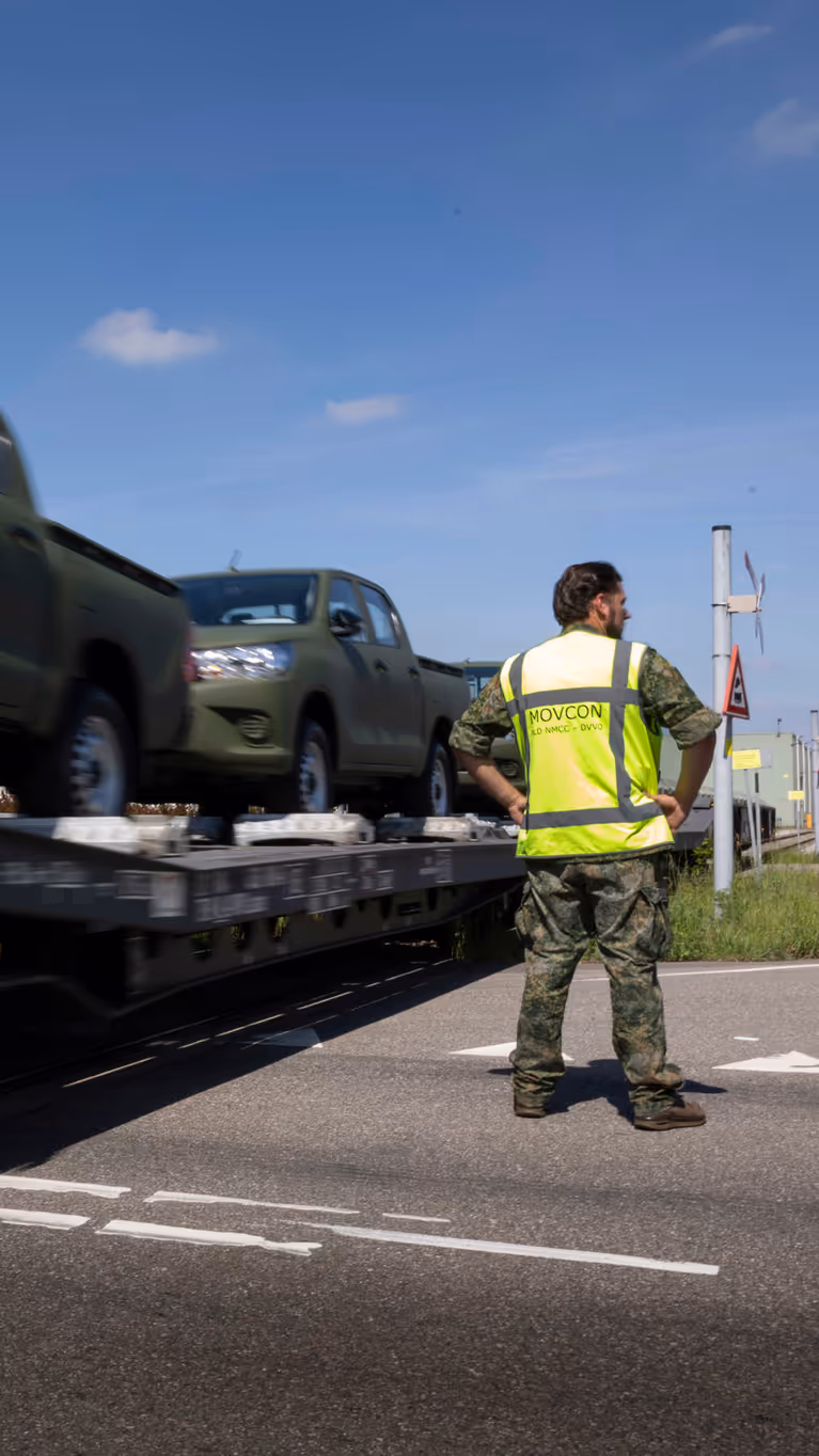 Een militair in geel hesje staat bij een voorbijrijdende trein met daarop voertuigen. 