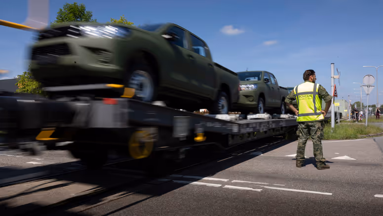 Een militair in geel hesje staat bij een voorbijrijdende trein met daarop voertuigen. 