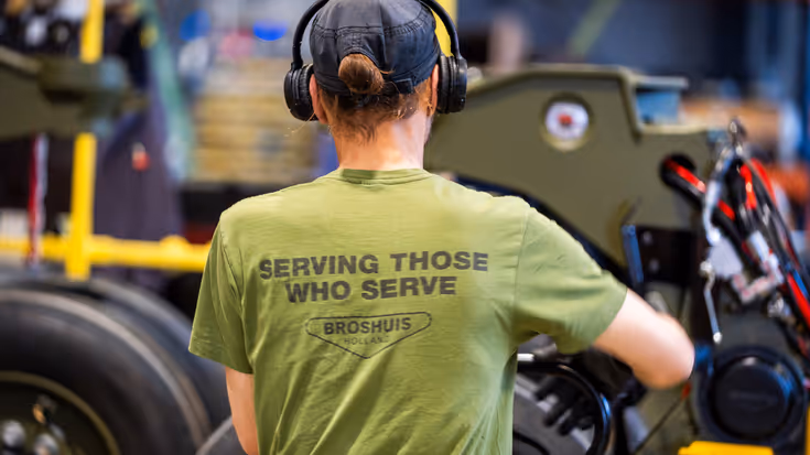 Monteur sleutelt aan wiel in garage en draagt T-shirt met tekst ‘serving those who serve’ op rug.