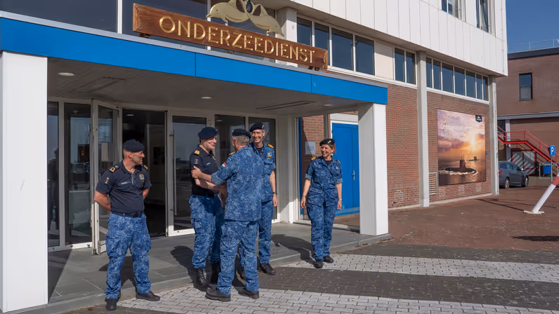 Een marineman schudt bij de ingang van een gebouw een collega de hand in de nabijheid van 3 andere marinemensen. 
