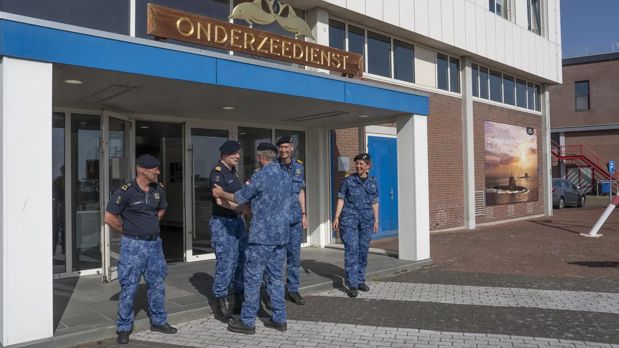 Een marineman schudt bij de ingang van een gebouw een collega de hand in de nabijheid van 3 andere marinemensen. 