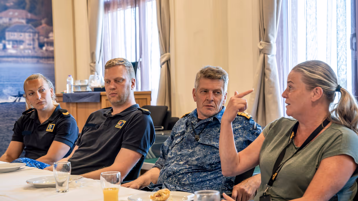 3 marinemensen luisteren aandachtig naar een vrouw in burgerkleding tijdens een lunch die op tafel staat.