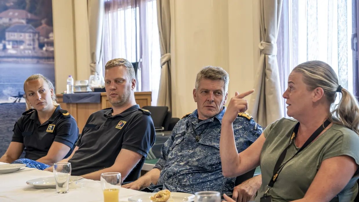3 marinemensen luisteren aandachtig naar een vrouw in burgerkleding tijdens een lunch die op tafel staat.