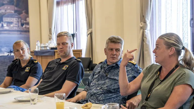 3 marinemensen luisteren aandachtig naar een vrouw in burgerkleding tijdens een lunch die op tafel staat.