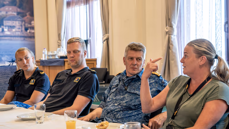 3 marinemensen luisteren aandachtig naar een vrouw in burgerkleding tijdens een lunch die op tafel staat.