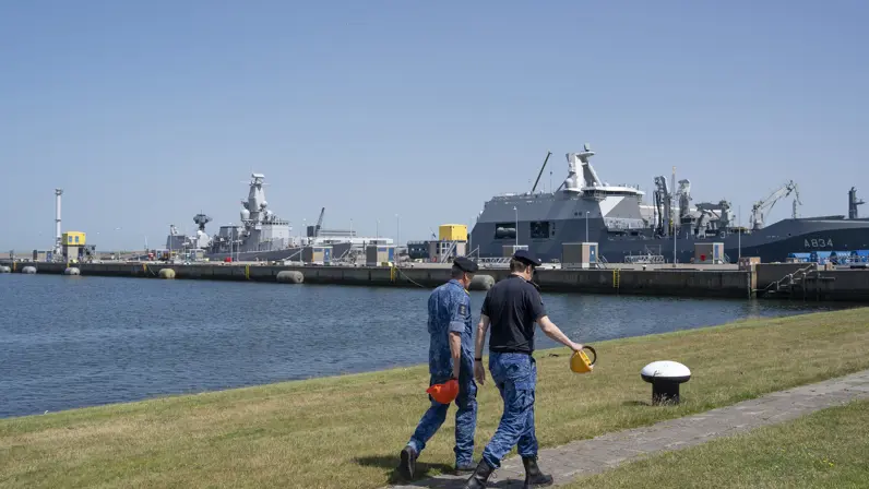 2 marinemensen lopen zij aan zij op de kade met op de achtergrond 2 marineschepen. 
