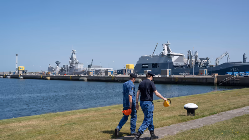 2 marinemensen lopen zij aan zij op de kade met op de achtergrond 2 marineschepen. 