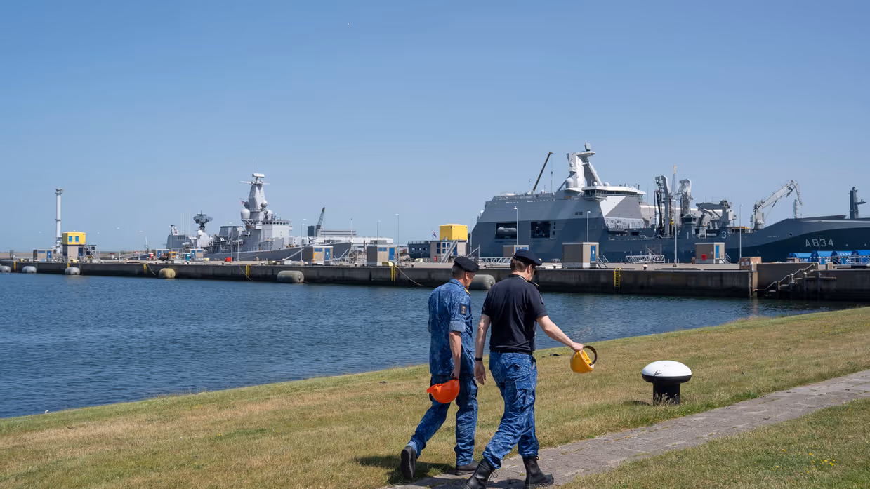 2 marinemensen lopen zij aan zij op de kade met op de achtergrond 2 marineschepen. 
