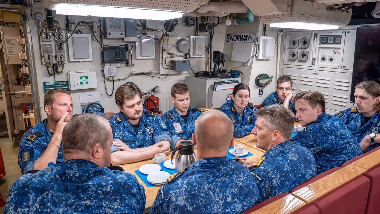 10 marinemensen praten met elkaar aan een tafel in een onderzeeboot.&nbsp;