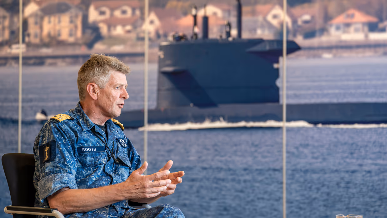 Vice-admiraal Boudewijn Boots praat met beide handen vanuit zijn stoel, voor een poster van een onderzeeboot. 