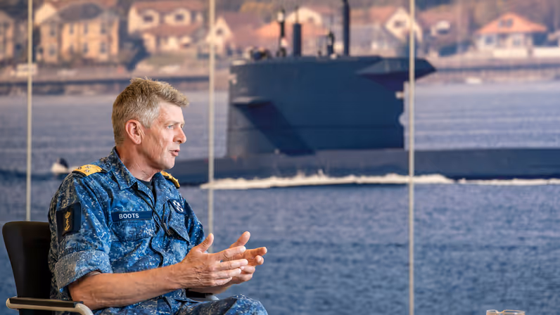 Vice-admiraal Boudewijn Boots praat met beide handen vanuit zijn stoel, voor een poster van een onderzeeboot. 