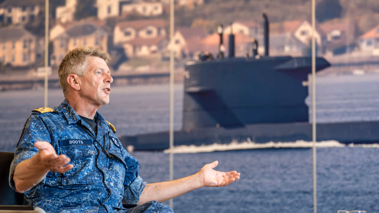 Een viceadmiraal praat met gespreide armen en open handen met op de achtergrond een foto van een onderzeeboot.