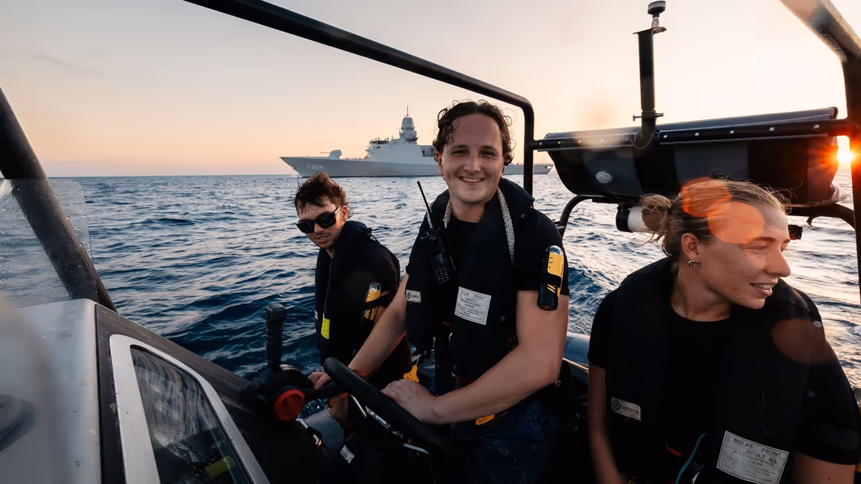 3 marinemensen varen met een RHIB, op de achtergrond ligt Zr.Ms. De Ruyter op zee.