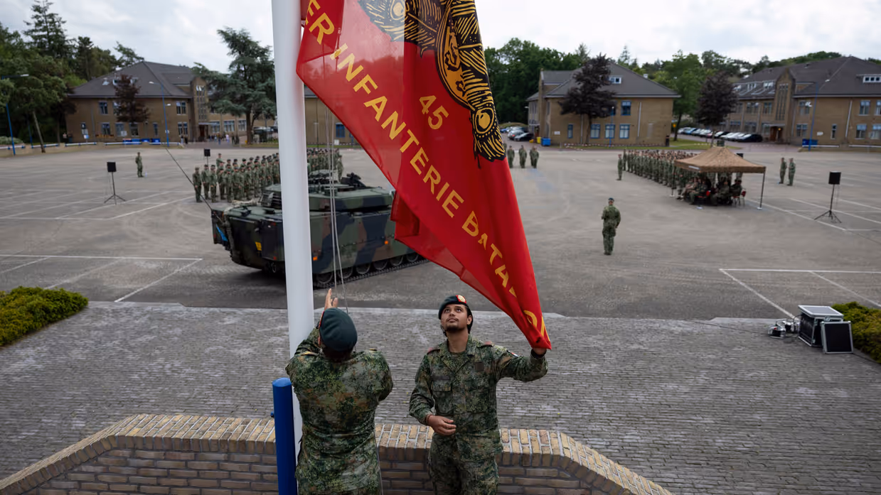 Twee militairen in camouflage hijsen een rode vlag met gouden tekst en embleem op een plein met militaire voertuigen en een opgestelde groep soldaten.