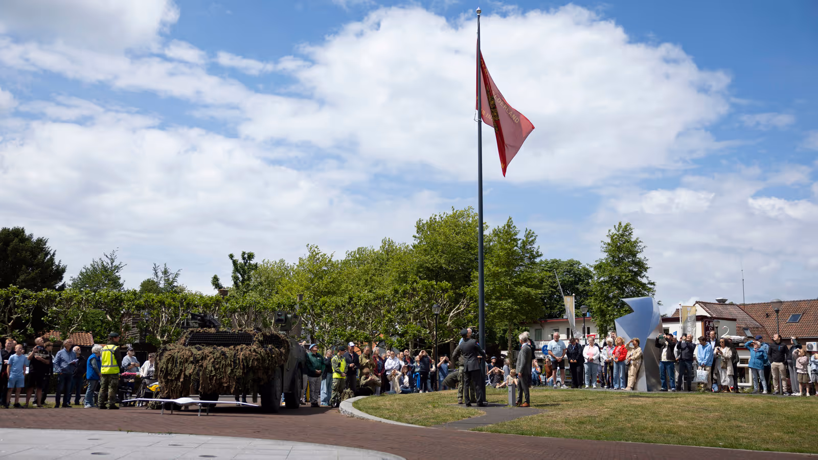 Een groep mensen kijkt naar een gecamoufleerde pantserwagen met een rode vlag op een zonnig plein met bomen en huizen.